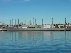 A small warship tied up alongside at a wharf. Her communications and radar masts have been crudely downsized, she carries no weapons, and several large squares have been cut into the ship's hull.