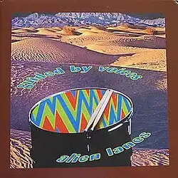 A desert landscape with a colorful drum in the foreground. The band name and album title encircle the drum like Saturn’s rings.