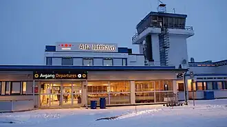 The entrance to a small airport terminal, mostly with one story, but also with a control tower to the right and a second story in the middle. On the second story is a sign with "Avinor" and "Alta lufthavn", while above the door it says "Departures"