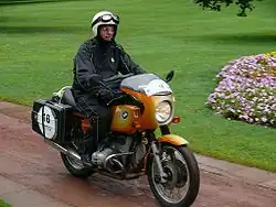 Gold BMW R90S motorcycle ridden through a park by a rider in black waterproof one piece suite