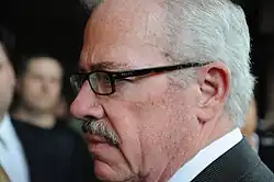 Left side, close up of a gray-haired mustached man with thick rimmed Prada glasses