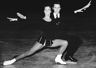 An image of Annerose Baier and Eberhard Rüger skating together. Both do a similar pose where they have their hands extended, doing a lunge and facing the camera.