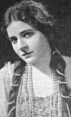 A young white woman wearing her long dark hair parted center and in long braids wrapped in pearls, with further long strands of pearls around her neck