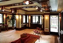 The western end of the living room. There is a protruding niche with windows at the end of the living room. In addition, there are lamps on the ceiling and walls, as well as a red sofa in the foreground.