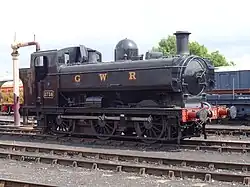 A black pannier tank locomotive, with the letters GWR in yellow on the side, pulls a mixed traffic train. The train includes a blue tank wagon, with "Express Dairy Milk for London" written on the side, and then a passenger carriage painted cream and red.