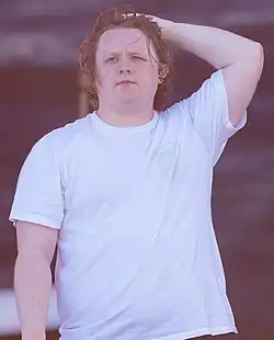 A man with shoulder-length brunette hair wearing a plain white t-shirt on a stage, one arm raised with the hand atop his head, the other arm down by his side. An earpiece is visible in his left ear.