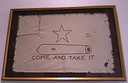 A white flag, with a five-pointed-star over the outline of a small cannon, with the words Come and Take It below. The left corner of the flag has been torn off and sewn back on.