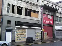 Grey stone-fronted building with blackened empty windows and shutters down. Graffiti reads "Who burnt us out? People before profit" - and "Fat cats get fatter, puts the kittens out in the cold."