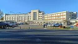 a large brick and concrete building stretches out across the photo about 6 stories high, cars and car parks and bare trees in the front. the sky is blue.