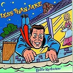 A man in a blue sit jumping off a diving board into a swimming pool as a woman from the house watches.