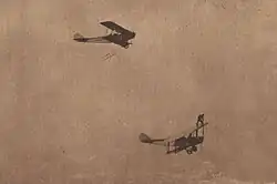 Two biplanes flying, with Ormer Locklear standing on the top wing of the lower airplane