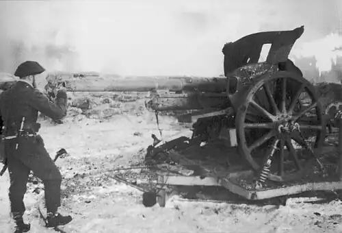 Black-and-white photograph of Churchill in uniform looking down the barrel of a large artillery gun with A stone barricade visible in the background