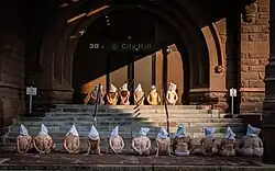 Seventeen naked people seated on ground in front of City Hall with their backs to the camera.