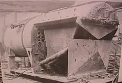 A picture of a steam locomotive boiler under construction. The firebox is a cube with curved top and is in pieces ready for welding. Various components are scattered about the floor of a boilershop at a locomotive works site.