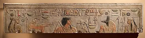 Light gray stone surface with carved and painted images of two woman, a falcon-headed god, a black-haired man with a long goatee, a jackal-headed god, and Egyptian hieroglyphs inscribed along the top