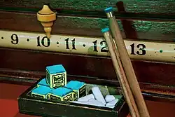Close-up view of a horizontal scoring band showing the numerals 9, 10, 11, 12, and 13, with a ceramic pointer resting above the 10, two cue tips resting against the 12, and a box of chalk sitting on a shelf beneath