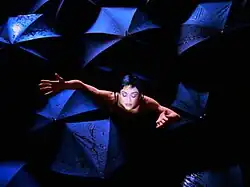 A black-haired woman with closed eyes and open arms stands among open dark blue umbrellas.