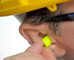 A picture of a man's head, focused on the ear, with yellow ear plugs being inserted into that ear.