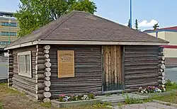 A small one-story square brown building of logs with the ends unfinished at the corners. It has a peaked roof with brown shingles and yellow trim on top. In front are flower beds; it has a yellow sign with fancy text headed "Yellowknife's Original School" at left and a wooden door of vertical boards in the middle. Behind it is a tree and part of two larger modern buildings.
