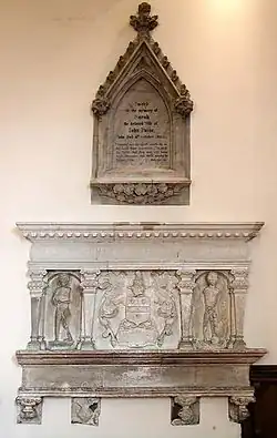 Two elaborate stone tablets set into a plain wall and lit from above. One is pentagonal and has black writing in an arched recess; the other, immediately below, is larger, rectangular and has effigies of three people.