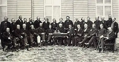 A photograph of a group of men in a semi-circle consisting of two rows. The front row is seated. There is a desk in the middle of the semi-circle with a man seated behind it.