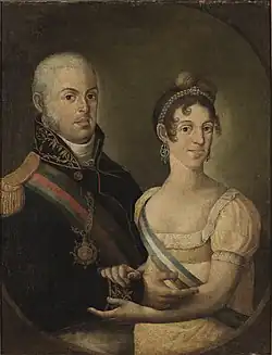Half-length portrait of a royal couple facing each other, hands clasped and looking ahead. He has light-coloured hair and wears a dark military uniform. She wears pearls, dangling earrings, and a white dress with a blue and white sash.
