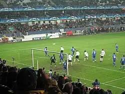 White and blue players standing inside the penalty area during a corner kick