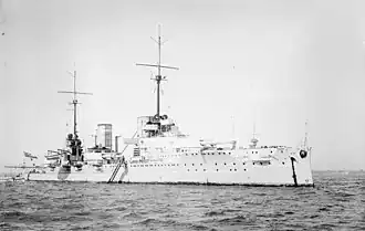 A large light gray warship sits motionless in harbor. Two tall masts stand in the center of the ship.