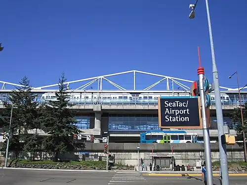 Elevated train station seen above a freeway on an embankment. A bus stop can be seen below the embankment and across a road from a sign reading "SeaTac/Airport Station" in the foreground.
