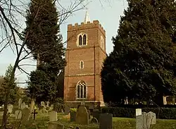 A brick tower with a battlemented parapet and a spirelet seen between trees