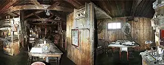 Dimly lit interior of a timber structure with a long table on which are papers and other unidentifiable objects. There are also bunks, sheles and packing cases. To the right, under a small window, is a smaller table with a chair.