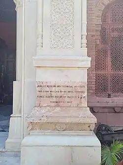 This is a rectangular stone plaque with inscription about laying the cornerstone for Museum - as Jubilee Museum and Technical Institute - by Prince Albert Victor February 3rd 1890. The stone is light in colour, the inscription is in English, in four rows, with black letters. This plaque is built in the decorative pillar at the right side of the Museum entrance.