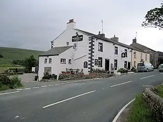 A largely whitewashed building with upland behind, and a road in front
