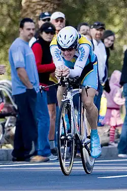 A cyclist wearing a blue and white jersey with yellow trim crouched into an aerodynamic position on his bicycle, riding down a road with spectators on the side watching him