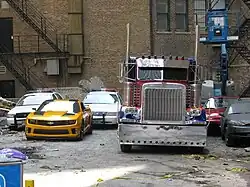 A Peterbilt 379, and a yellow Camaro are beside each other, with two police cars behind. They are near a building complex.