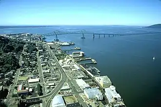 A long bridge crosses a huge river flowing into a vast body of water under a blue sky. The bridge begins in a settlement with streets, buildings, and wharves along the river and extends out of sight toward a low hill on the far shore. The first part of the bridge has a superstructure and is high above the water but then the bridge gradually descends and continues out of sight not so far above the water.