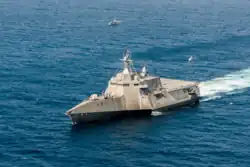 A U.S. Navy ship at sea with a manned helicopter flying overhead and an unmanned helicopter flying above her stern.