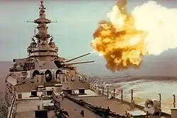 A large gunship facing the viewer with one gun turret pointed toward the right. Flames can be seen from the gun barrels, and a concussive effect is visible on the water.