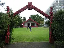 A Māori carved gate way frames a Māori meeting house with two active young people in between on the grass area.