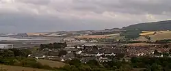 View of multiple houses with sea on the left and hills in the background.
