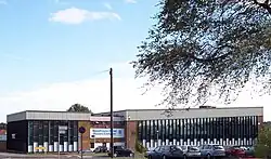A building approximately 3 stories high. For the most part, the building is made up of horizontal columns of an opaque white panel up to around head height, with 3 windows arranged vertically above this. The columns are approximately 0.5&nbsp;m (1.6&nbsp;ft) wide and run from just below the roof to just above ground level and are separated by vertical wooden beams. Above the columns of windows a corrugated white border runs along the top of the building.