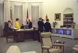 several people standing and sitting around the Johnson Desk with three television screens to the right displaying news programs.